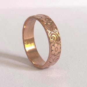 May include: A copper-colored ring with a floral pattern. The ring features an embossed design of flowers and swirling leaves around the band. The ring is a simple band style.