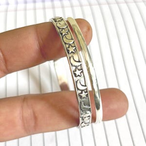May include: Two silver bangles. One bangle features a repeating pattern of stars and crescent moons. The other bangle is a plain, polished silver band. The bangles are displayed against a white background.