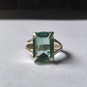May include: A silver ring featuring a large, rectangular, teal-colored gemstone. The gemstone is set in a silver band with a split shank design. The ring is displayed on a white surface.