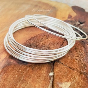 Puede incluir: Una pila de finas pulseras plateadas, que reflejan la luz, están dispuestas sobre una superficie de madera marrón texturizada. Las pulseras son circulares y parecen ser de metal. La imagen está bien iluminada, resaltando el brillo de las joyas.