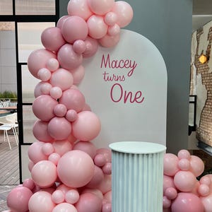 May include: A display for a first birthday celebration, featuring a pink balloon arch and clusters. The backdrop is white with the text "Macey turns One." A small, white, fluted pedestal table is in front. The venue has outdoor seating.