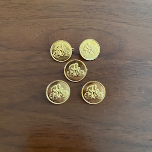 May include: Five gold-colored round buttons with a cyclist design. Each button features a detailed relief of a person riding a bicycle. The buttons are arranged on a dark wooden surface.