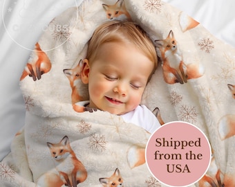 May include: A cream-colored baby blanket with a repeating pattern of orange foxes and snowflakes. A sleeping child is wrapped in the blanket. The blanket has a soft, plush texture, perfect for a baby.