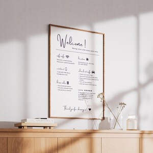 May include: A framed welcome sign with a wooden frame, featuring handwritten text in dark blue. The sign includes information about Wi-Fi, contact details, house rules, and check-out instructions. The sign is displayed on a wooden cabinet.
