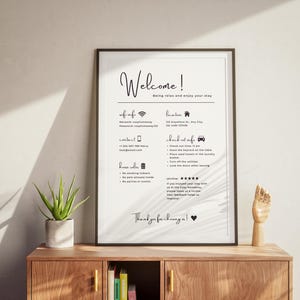 Framed welcome sign on a wooden shelf details Wi-Fi info, location, contact, and house rules. Nearby are a potted plant and a wooden hand model. Warm, inviting tone.