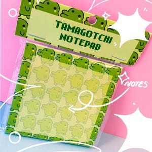 May include: A Tamagotchi notepad in a clear plastic package. The notepad features a repeating pattern of yellow cartoon characters on a light yellow background. The top of the notepad has the words "TAMAGOTCHI NOTEPAD" in green.