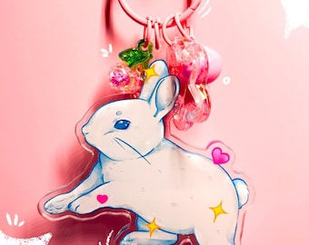 Könnte beinhalten: Ein rosa Schlüsselanhänger mit einer weißen Kaninchen-Illustration mit blauen Akzenten, Herzen und Sternen. Der Schlüsselanhänger enthält einen rosa Ring und mehrere Anhänger, vor einem rosa Hintergrund.