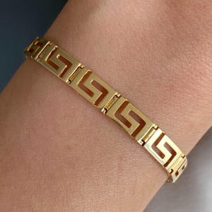 May include: A gold-toned bracelet featuring a repeating geometric Greek key pattern. The metal links are connected in a continuous band design. The text Adelaide Jewels is printed in a small black box in the bottom left corner of the image.