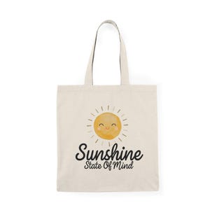 Puede incluir: Bolso tote color crema con un gráfico de sol sonriente y el texto "Sunshine State Of Mind" en escritura negra. El sol es amarillo con líneas radiantes. El bolso tiene dos asas largas.