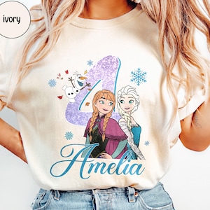 May include: Ivory-colored t-shirt featuring a graphic of two animated princesses, a purple number 4, snowflakes, and the name "Amelia" in a script font. The design includes a cartoon snowman and other decorative elements.