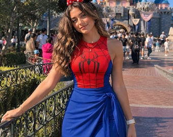 May include: A sleeveless dress featuring a red bodice with a black spiderweb pattern and a black spider emblem, paired with a solid blue skirt that has a side tie detail. The dress is worn with black mouse ears featuring a red and white polka dot bow.