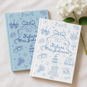 May include: Two hardback journals, one pale blue and one white, with blue illustrations. The journals feature a bow, rings, and the text "Future Mrs. Johnson". Decorative elements include a cake, champagne glasses, and fireworks.