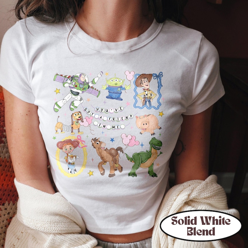 May include: A white short-sleeve t-shirt featuring a graphic print of Toy Story characters including Buzz Lightyear, Woody, Jessie, Rex, Slinky Dog, Hamm, Bullseye, and an alien. The text "YOU'VE GOT A FRIEND IN ME" is printed in the center.