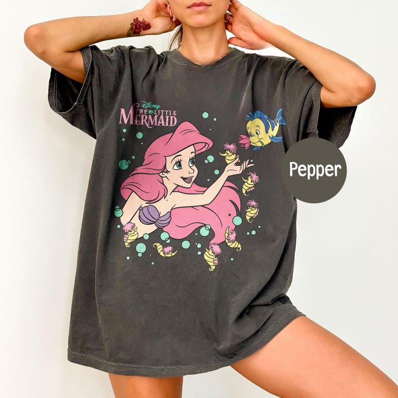 May include: A charcoal gray oversized t-shirt featuring a graphic print of Ariel from The Little Mermaid with pink hair, surrounded by bubbles and small sea creatures. The text The Little Mermaid is printed above the character, and the word Pepper is visible.