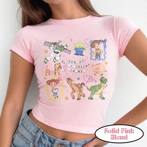 May include: A light pink short-sleeved t-shirt featuring a graphic print of various animated characters including a space ranger, a green alien, a cowboy, a cowgirl, a spring dog, a pig, a horse, and a dinosaur with the text You've Got A Friend In Me.