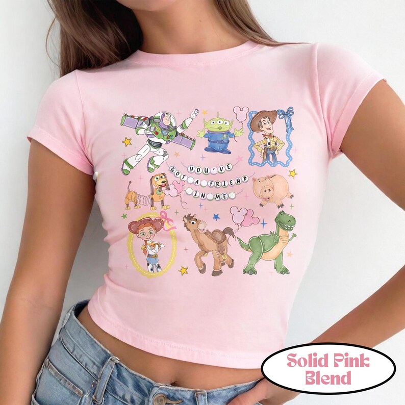 May include: A light pink short-sleeved t-shirt featuring a graphic print of various animated characters including a space ranger, a green alien, a cowboy, a cowgirl, a spring dog, a pig, a horse, and a dinosaur with the text You've Got A Friend In Me.