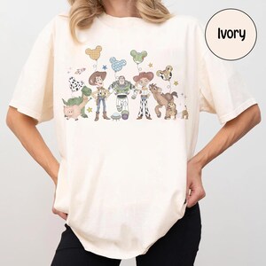May include: Ivory-colored t-shirt featuring a colorful illustration of animated characters, including a dinosaur, cowboy, and space ranger. The graphic includes balloons and stars. The word "Ivory" is printed on a circular label.