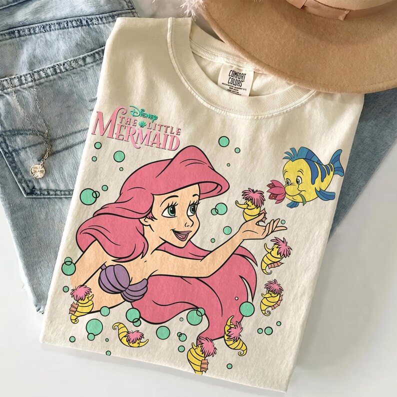 May include: A cream-colored t-shirt featuring a graphic of Ariel from The Little Mermaid with long pink hair, a purple seashell top, and a yellow fish. The design includes small seahorses, green bubbles, and the text Disney The Little Mermaid.