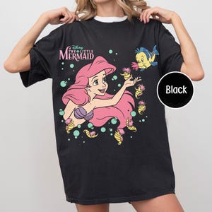 May include: A black short-sleeve t-shirt featuring a graphic of Ariel from Disney's The Little Mermaid with long pink hair, a purple seashell top, and several small yellow sea creatures. The text Disney The Little Mermaid is printed above.
