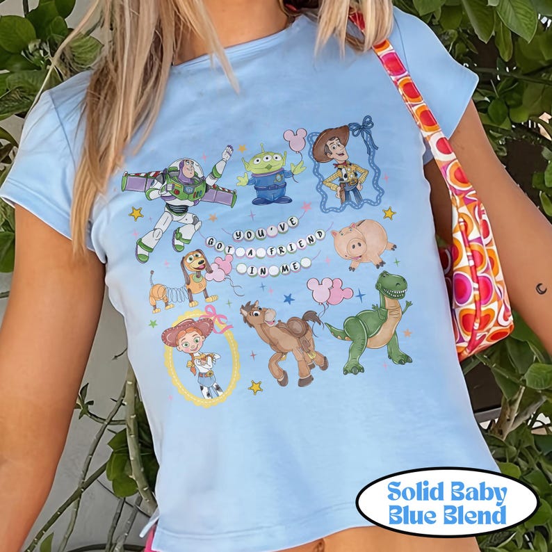 May include: A light blue t-shirt featuring a graphic print of Toy Story characters including Buzz Lightyear, Woody, Jessie, Rex, Slinky Dog, Hamm, Bullseye, and an alien. The text "YOU'VE GOT A FRIEND IN ME" is printed in the center with small stars.