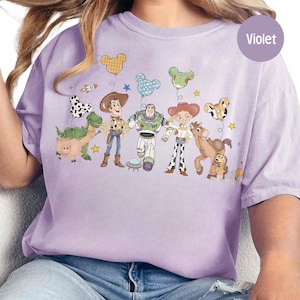 May include: A light purple t-shirt featuring a graphic of Toy Story characters with Mickey Mouse-shaped balloons. The design includes Woody, Buzz Lightyear, Jessie, and other characters. The shirt has a relaxed fit and a round neckline.