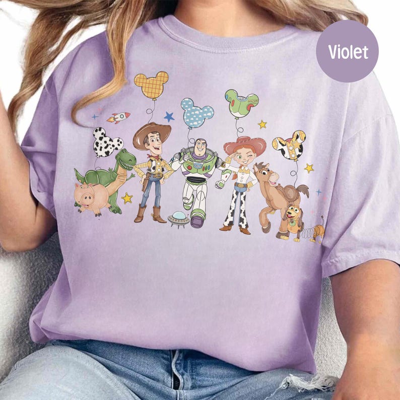 May include: A light purple t-shirt featuring a graphic of Toy Story characters with Mickey Mouse-shaped balloons. The design includes Woody, Buzz Lightyear, Jessie, and other characters. The shirt has a relaxed fit and a round neckline.
