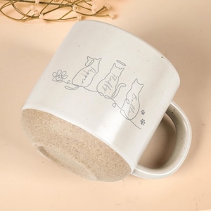 May include: Cream-colored ceramic mug with a light brown base and a white handle. The mug features line drawings of a dog and two cats, with the names "Pepper", "Fluffy", and "Bella" written below each animal. Paw prints and a flower are also included.