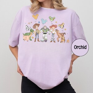 May include: A light purple t-shirt featuring a cartoon graphic of characters from a popular animated movie. The graphic includes characters like a cowboy, a space ranger, and a dinosaur, with balloons and stars. The word "Orchid" is in a circle.