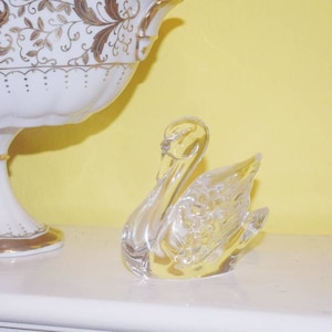 May include: A white porcelain bowl with gold floral designs sits on a white wooden cabinet. A clear glass swan figurine is placed on the cabinet in front of the bowl.