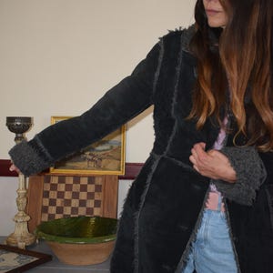 Può includere: Un lungo cappotto nero con finiture in ecopelliccia. Il cappotto è aperto, rivelando un top rosa e jeans blu. Il cappotto ha un aspetto strutturato e un design classico. Le maniche hanno finiture in ecopelliccia.