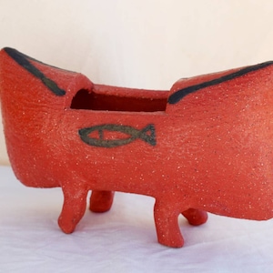 May include: Red ceramic planter shaped like a boat with four legs and a black fish design.