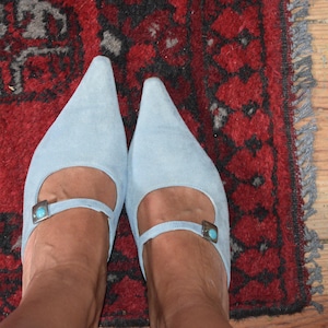 Puede incluir: Un par de zapatos de ante azul claro con punta, correas en el tobillo y pequeñas hebillas plateadas. Los zapatos están sobre una alfombra roja con un patrón oscuro y suelo de madera.