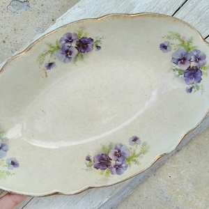 以下が含まれることがあります： 白い背景と金色の縁取りのある楕円形のサービングディッシュ。ディッシュは紫と緑の花柄で飾られています。