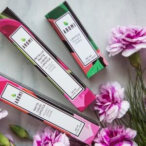 May include: Three pink and green lipstick boxes with the brand name "AROMI" and the text "Metallic Matte Liquid Lipstick" and "Matte Liquid Lipstick" printed on the boxes. The boxes are arranged on a white marble surface with pink carnations.