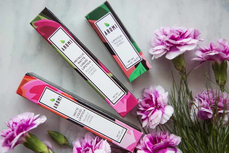 May include: Three pink and green lipstick boxes with the brand name "AROMI" and the text "Metallic Matte Liquid Lipstick" and "Matte Liquid Lipstick" printed on the boxes. The boxes are arranged on a white marble surface with pink carnations.