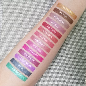 K&ouml;nnte beinhalten: Eine Nahaufnahme eines Arms mit 14 Lidschatten-Swatches. Die Swatches sind vertikal von oben nach unten angeordnet. Die Lidschattenfarben sind Gold, Braun, Lila, Rot, Rosa und Gr&uuml;n. Die Namen der Lidschattenfarben sind unter jedem Swatch aufgeschrieben. Die Namen der Lidschattenfarben sind: Gold Digger, Teddy Bear, Brown Sugar, Pixie Dust, Candy Apple, Copper Bliss, Rose Gold, Pumpkin Spice, Pink Sugar, Fab Flamingo, Sugared Mauve, Purple Unicorn, Ice Age und Mermaid Tail.