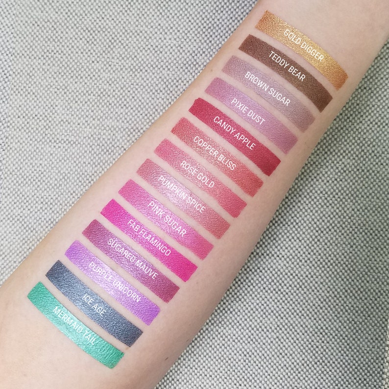 K&ouml;nnte beinhalten: Eine Nahaufnahme eines Arms mit 14 Lidschatten-Swatches. Die Swatches sind vertikal von oben nach unten angeordnet. Die Lidschattenfarben sind Gold, Braun, Lila, Rot, Rosa und Gr&uuml;n. Die Namen der Lidschattenfarben sind unter jedem Swatch aufgeschrieben. Die Namen der Lidschattenfarben sind: Gold Digger, Teddy Bear, Brown Sugar, Pixie Dust, Candy Apple, Copper Bliss, Rose Gold, Pumpkin Spice, Pink Sugar, Fab Flamingo, Sugared Mauve, Purple Unicorn, Ice Age und Mermaid Tail.