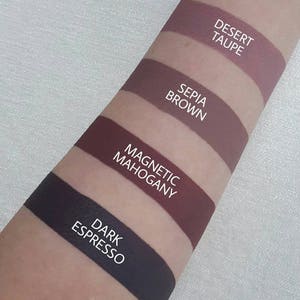 May include: Four different shades of lipstick swatches on a person's arm. The shades are labelled "Dark Espresso", "Magnetic Mahogany", "Sepia Brown", and "Desert Taupe".