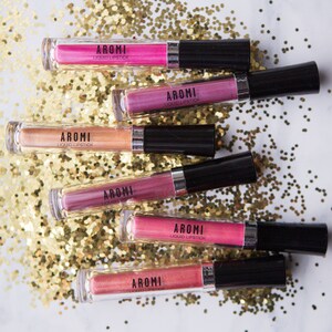 May include: Six tubes of AROMI liquid lipstick in various shades of pink, gold, and purple. The tubes are arranged on a bed of gold glitter.