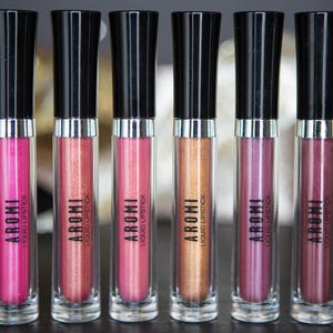 K&ouml;nnte beinhalten: Sechs transparente R&ouml;hrchen mit fl&uuml;ssigem Lippenstift, schwarzen Kappen und silbernen B&auml;ndern. Die R&ouml;hrchen sind mit "AROMI LIQUID LIPSTICK" beschriftet. Die Lippenstifte sind in Rosat&ouml;nen, Rot und Gold.