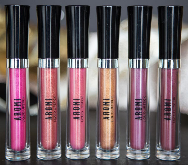 K&ouml;nnte beinhalten: Sechs transparente R&ouml;hrchen mit fl&uuml;ssigem Lippenstift, schwarzen Kappen und silbernen B&auml;ndern. Die R&ouml;hrchen sind mit "AROMI LIQUID LIPSTICK" beschriftet. Die Lippenstifte sind in Rosat&ouml;nen, Rot und Gold.
