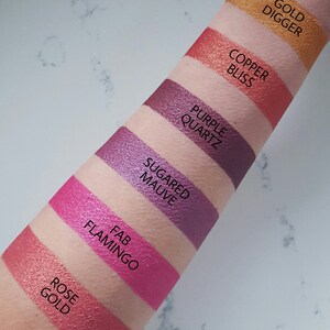 May include: A close-up of a person's arm with swatches of eyeshadow in various shades of pink, purple, and copper. The swatches are labeled with names like "Rose Gold", "Fab Flamingo", "Sugar Mauve", "Purple Quartz", "Copper Bliss", and "Gold Digger".