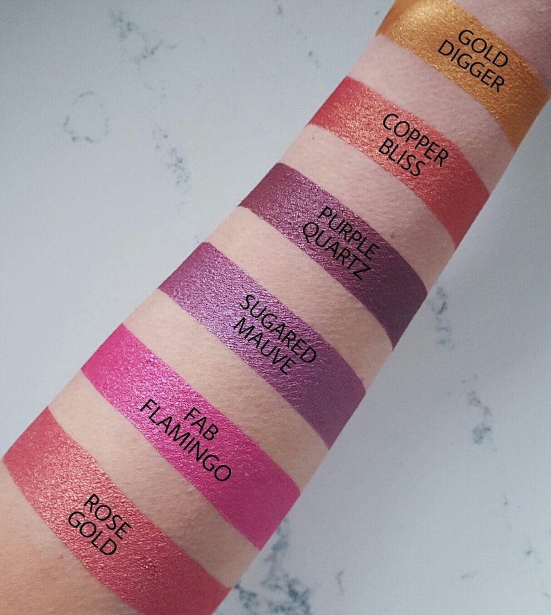 May include: A close-up of a person's arm with swatches of eyeshadow in various shades of pink, purple, and copper. The swatches are labeled with names like "Rose Gold", "Fab Flamingo", "Sugar Mauve", "Purple Quartz", "Copper Bliss", and "Gold Digger".