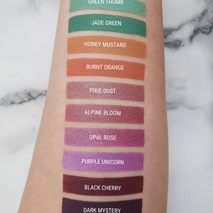 Puede incluir: Una muestra de 10 colores de sombra de ojos diferentes en el brazo de una persona. Los colores son: Green Thumb, Jade Green, Honey Mustard, Burnt Orange, Pixie Dust, Alpine Bloom, Opal Rose, Purple Unicorn, Black Cherry y Dark Mystery.