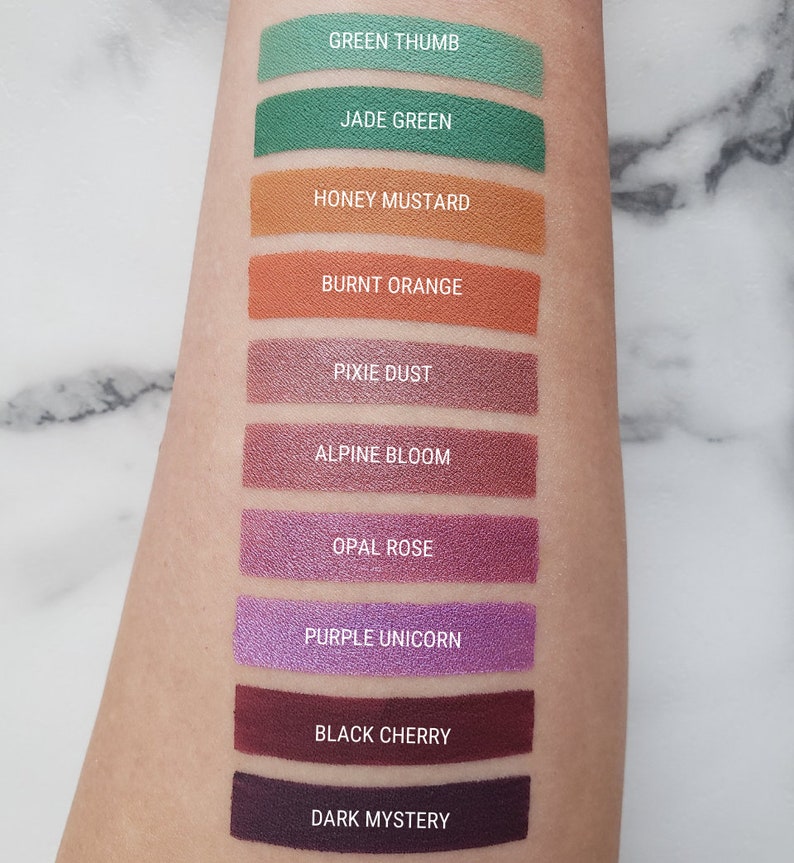 Puede incluir: Una muestra de 10 colores de sombra de ojos diferentes en el brazo de una persona. Los colores son: Green Thumb, Jade Green, Honey Mustard, Burnt Orange, Pixie Dust, Alpine Bloom, Opal Rose, Purple Unicorn, Black Cherry y Dark Mystery.
