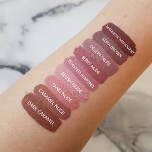 May include: A close-up of a person's arm with ten different shades of lipstick swatches. The shades are labelled from darkest to lightest: Magnetic Mahogany, Sepia Brown, Desert Taupe, Berry Nude, Toasted Almond, Blush Nude, Sand Nude, Caramel Nude, and Dark Caramel.