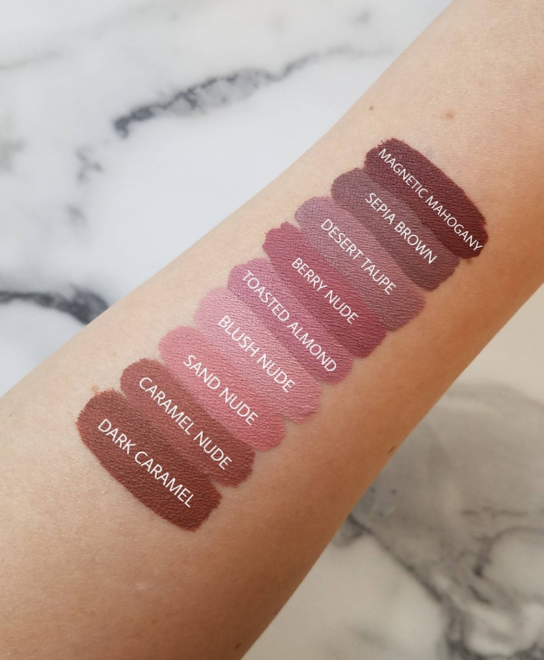 May include: A close-up of a person's arm with ten different shades of lipstick swatches. The shades are labelled from darkest to lightest: Magnetic Mahogany, Sepia Brown, Desert Taupe, Berry Nude, Toasted Almond, Blush Nude, Sand Nude, Caramel Nude, and Dark Caramel.