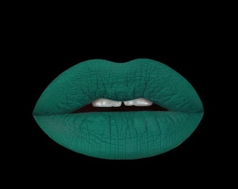 May include: A pair of lips coated in a matte, deep teal-green lipstick. The texture of the product appears dry and cracked across the surface of the lips, with a small portion of white teeth visible in the center gap between the upper and lower lip.
