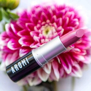 K&ouml;nnte beinhalten: Ein rosa Lippenstift mit silberner Kappe und schwarzer Basis. Auf dem Lippenstift-R&ouml;hrchen steht "AROMI". Der Lippenstift liegt auf einer rosa Blume.