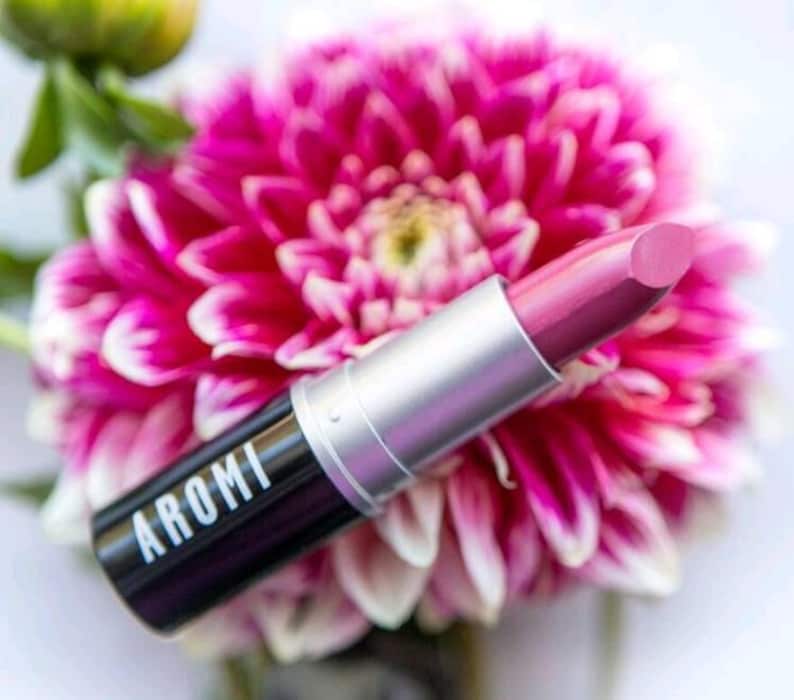 K&ouml;nnte beinhalten: Ein rosa Lippenstift mit silberner Kappe und schwarzer Basis. Auf dem Lippenstift-R&ouml;hrchen steht "AROMI". Der Lippenstift liegt auf einer rosa Blume.